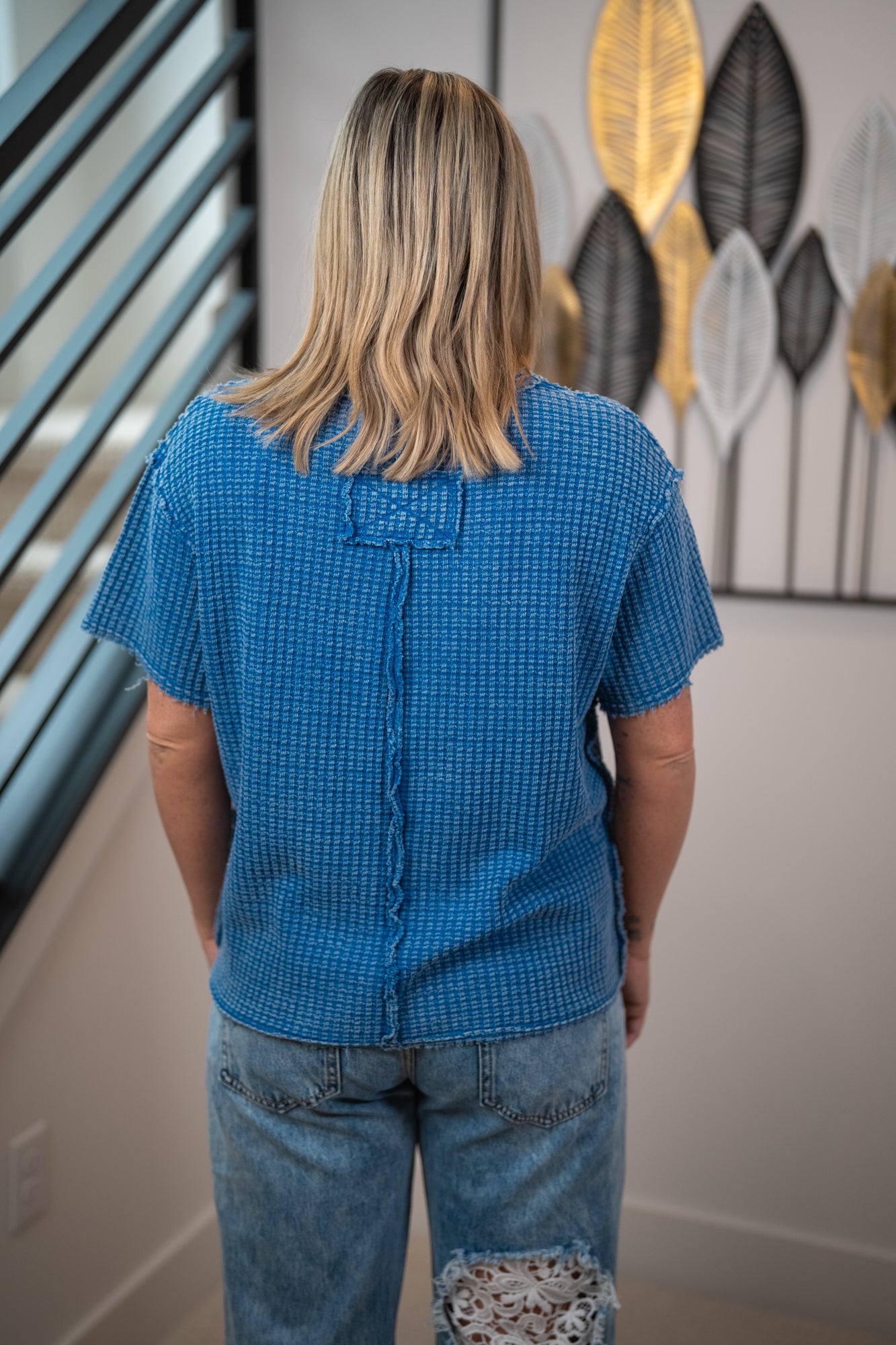 Person wearing a blue textured shirt and jeans standing in a room with decorative wall art.