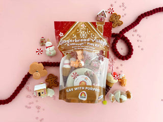 Gingerbread Village KidDough Play Kit bag surrounded by festive holiday figurines on pink background