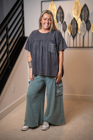 Woman wearing a dark gray oversized top and teal cargo pants standing in a room with decorative wall art.