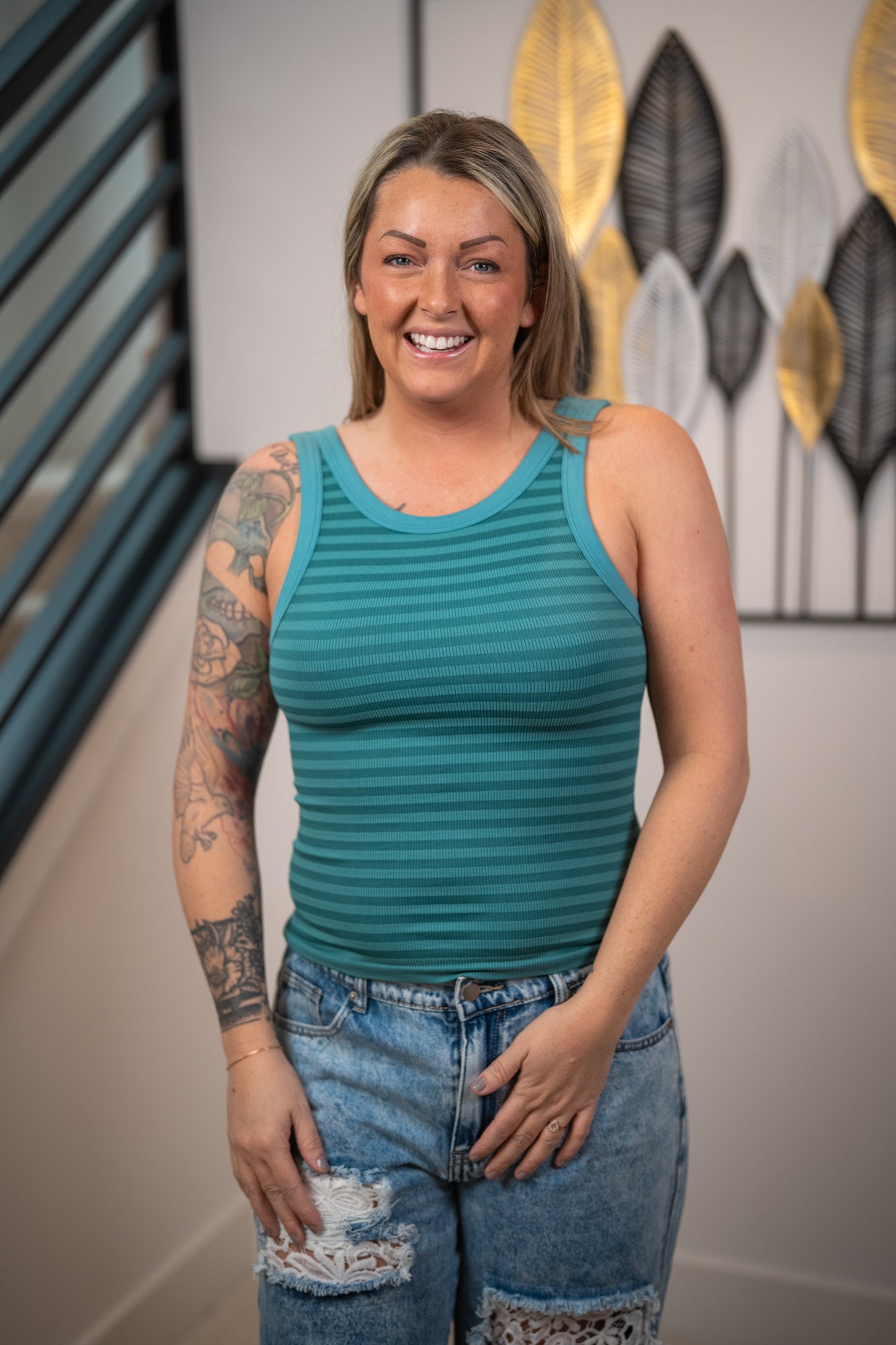 Woman wearing a teal tank top and denim shorts standing indoors with decorative wall art in the background.