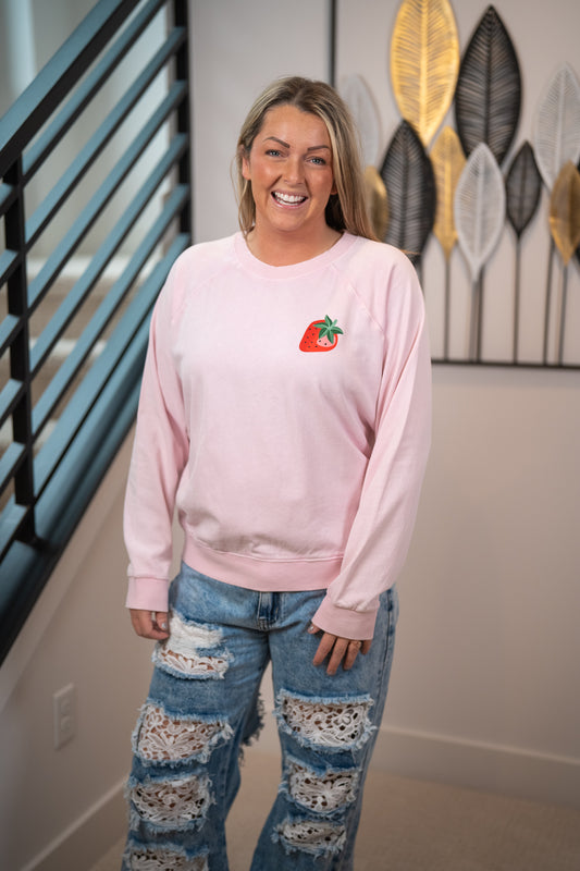 Person wearing a pink sweatshirt with a strawberry design and ripped jeans in a room.