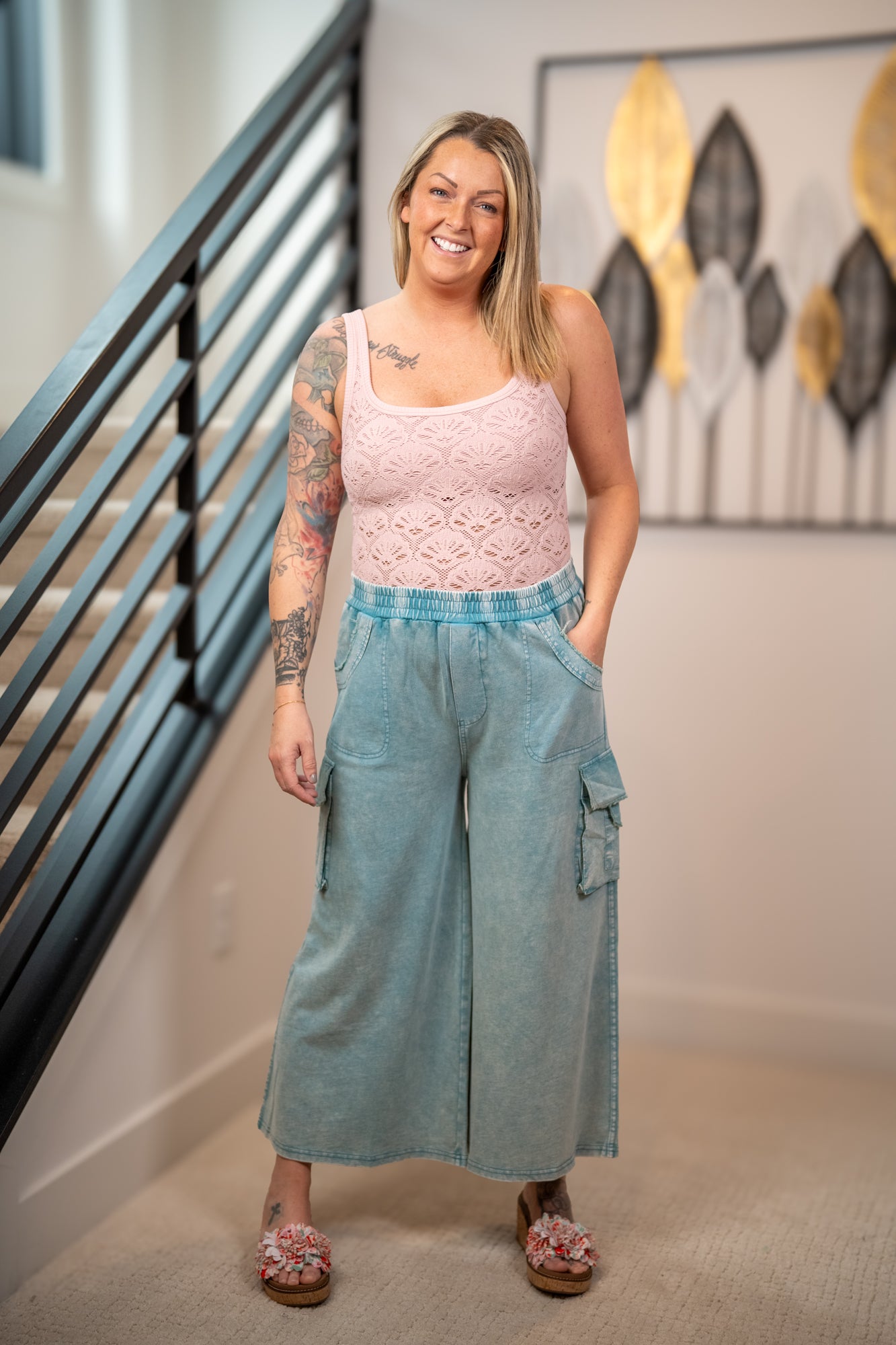Woman wearing a pink lace top and light blue wide-leg pants standing in a room with a staircase and decorative wall art.