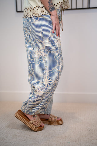 Person wearing blue striped pants with floral embroidery and sandals on a neutral background