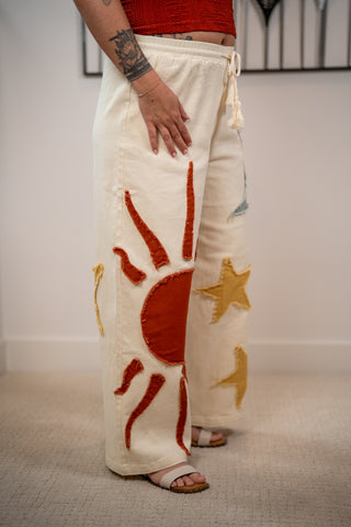 Person wearing cream-colored pants with red sun and yellow star designs, standing indoors.