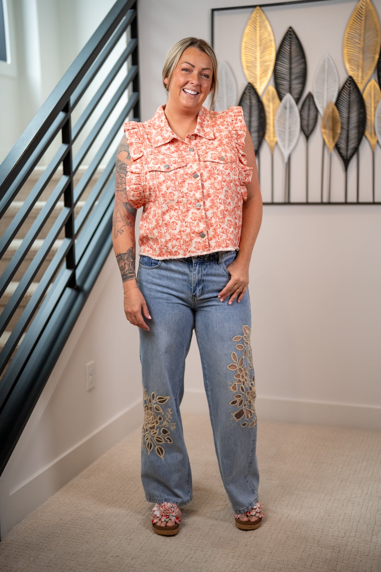 Woman wearing a floral top and jeans with embroidery, standing in a home setting.
