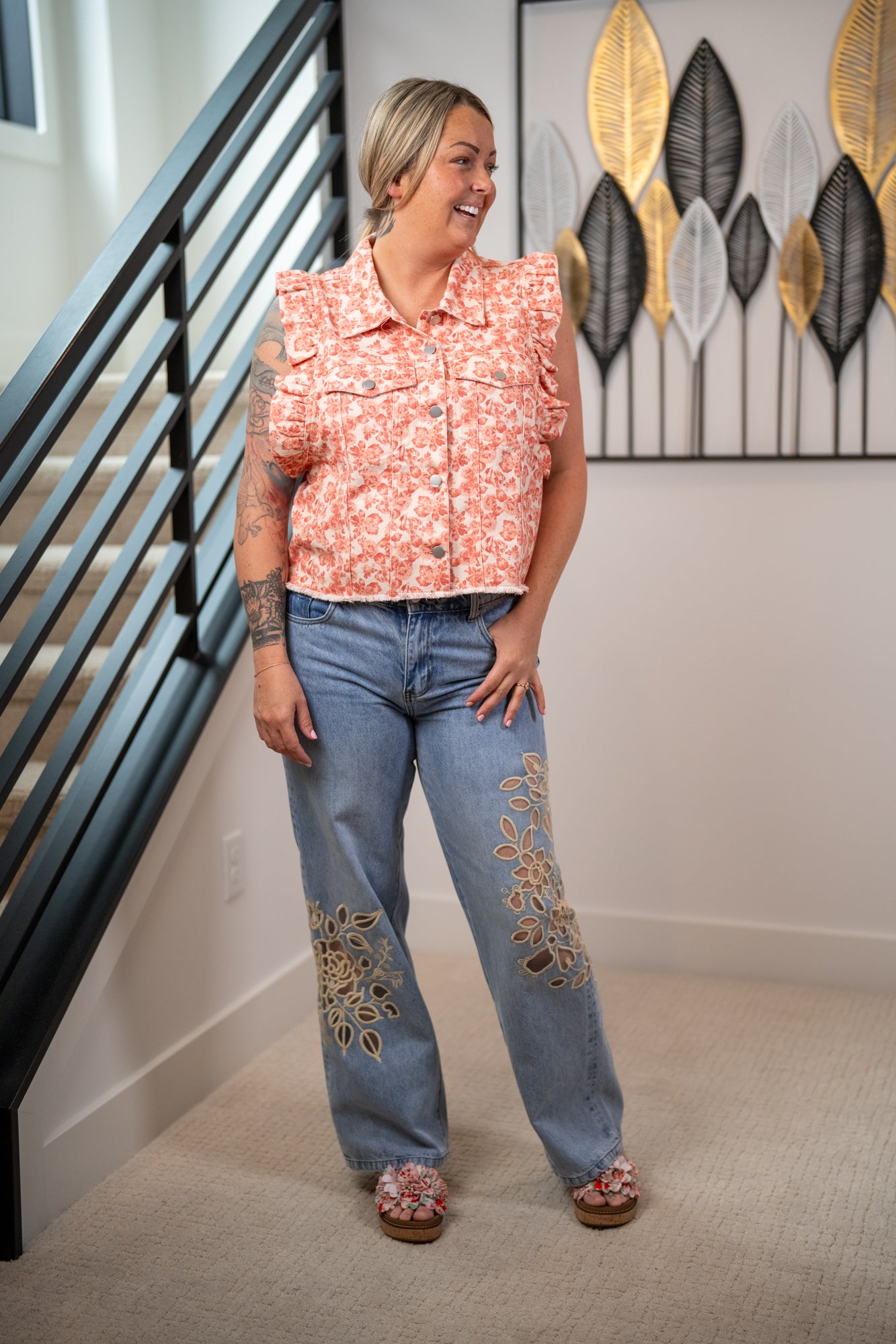 Woman wearing a floral top and jeans with embroidery, standing in a home setting.