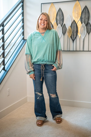 Woman wearing a teal top and blue jeans standing in a room with a decorative wall piece.
