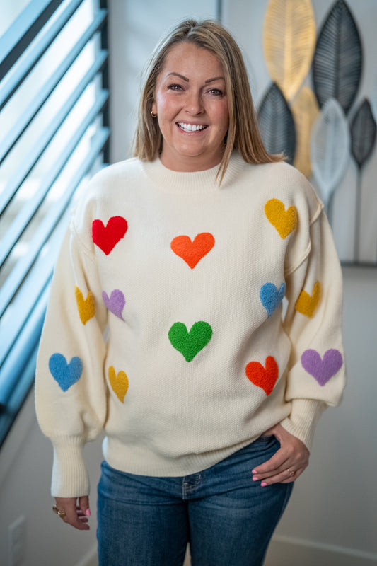 Woman wearing a cream sweater with colorful heart patterns indoors.