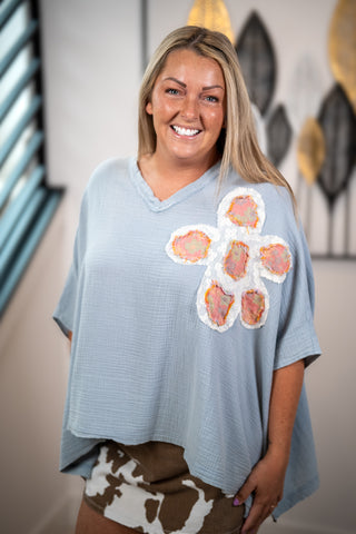A smiling blonde woman wearing a light blue waffle-knit poncho-style top with a large hand-painted flower applique on the chest, paired with brown and white cow-print shorts, standing in an indoor setting.