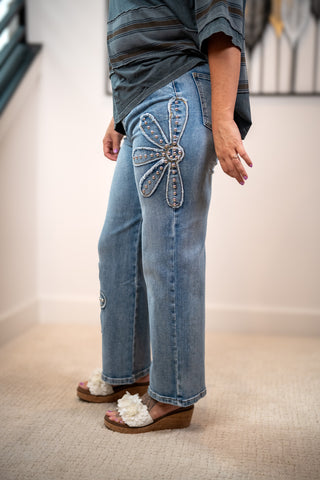 A person wearing light blue denim jeans with a large embroidered flower applique on the upper right leg, paired with a dark striped top and white fuzzy wedge sandals, standing indoors on a beige carpet.