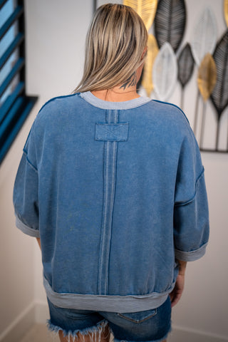 A woman viewed from behind wearing a washed blue cropped sweatshirt with a vertical seam detail down the back and ribbed cuffs, hem, and collar. She is also wearing frayed denim shorts in an indoor setting with a decorative wall hanging in the background.