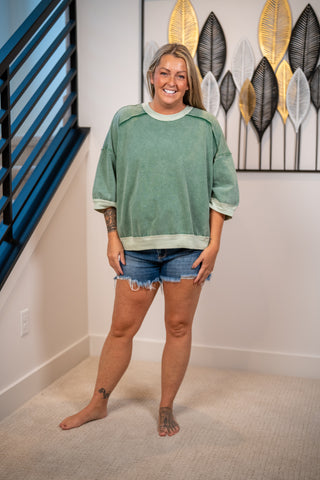 A woman stands barefoot in a home interior wearing an oversized sage green crewneck sweatshirt with rolled cuffs and denim cutoff shorts.