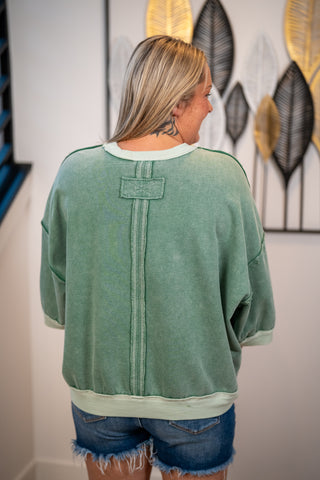 A woman viewed from behind wearing a sage green washed sweatshirt with a vertical seam detail on the back and denim shorts, standing indoors against a white wall with decorative wall art.