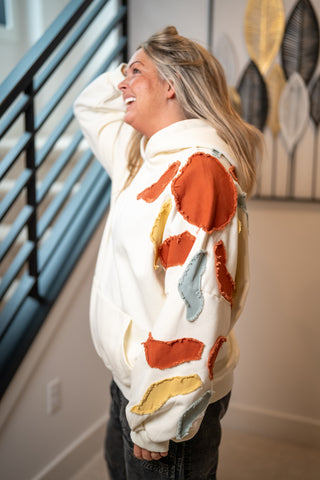 Cream hoodie with rust, blue, and yellow abstract fabric patches, kangaroo pocket