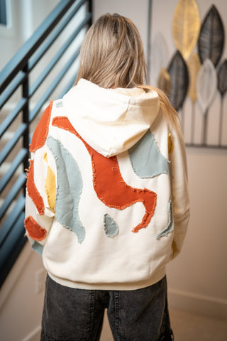 Cream hoodie with rust, blue, and yellow abstract fabric appliqué patches on back