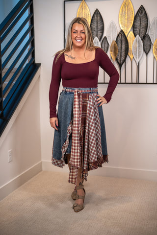 A woman modeling a unique outfit consisting of a maroon long-sleeve scoop-neck top, a denim skirt, and a brown and white plaid shirt tied around her waist, standing in an indoor setting with a staircase and wall art in the background.