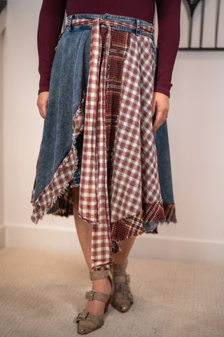 A person wearing a patchwork-style asymmetrical skirt made of blue denim and red-and-white plaid fabrics, paired with a burgundy long-sleeve top and taupe heels, standing indoors against a light wall.