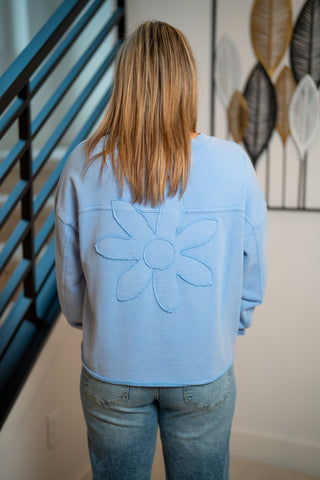 Person wearing a light blue sweatshirt with a floral design, standing in a room with a decorative wall art piece.