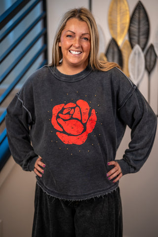 A smiling woman wearing a dark gray mineral-wash crewneck sweatshirt with a large red rose graphic on the front, paired with black pants, standing indoors against a wall with decorative leaf art.