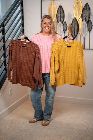 Mineral washed cable knit sweaters in brown, mustard, and pink on hangers