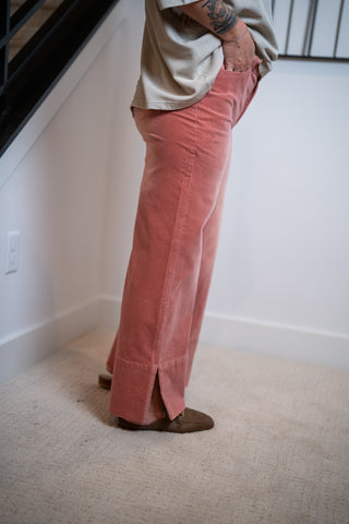 Person wearing pink corduroy pants and a beige shirt in a room with white walls and a carpeted floor.